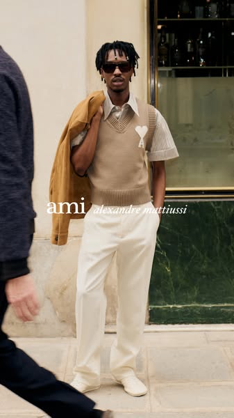 An image ad for AMI Paris features a Black male model wearing a tan sweater vest with a white heart and the AMI logo on 