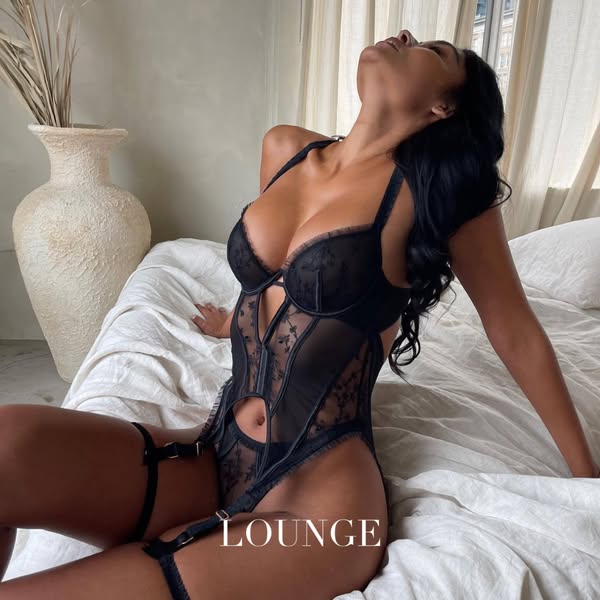 This ad features a woman in a black lingerie set from Lounge, posed on a bed in a bright room.