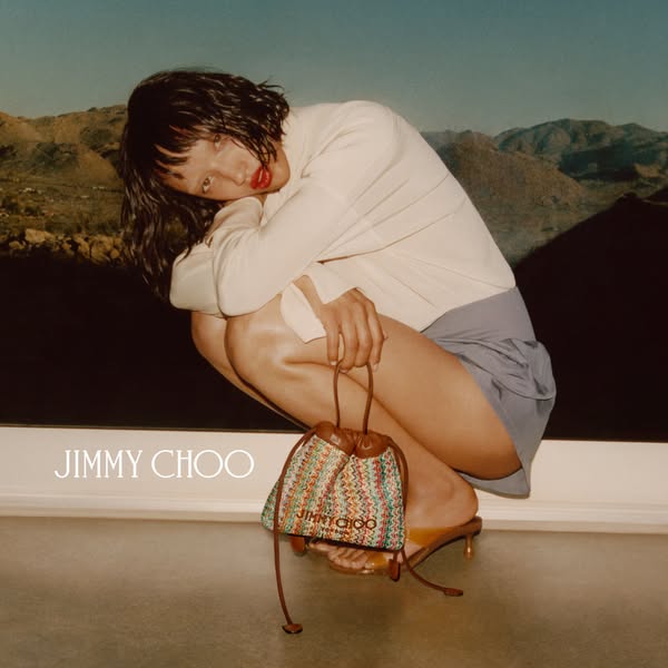 This ad features a woman in casual clothing, holding a Jimmy Choo bucket bag against a backdrop of a desert landscape. T