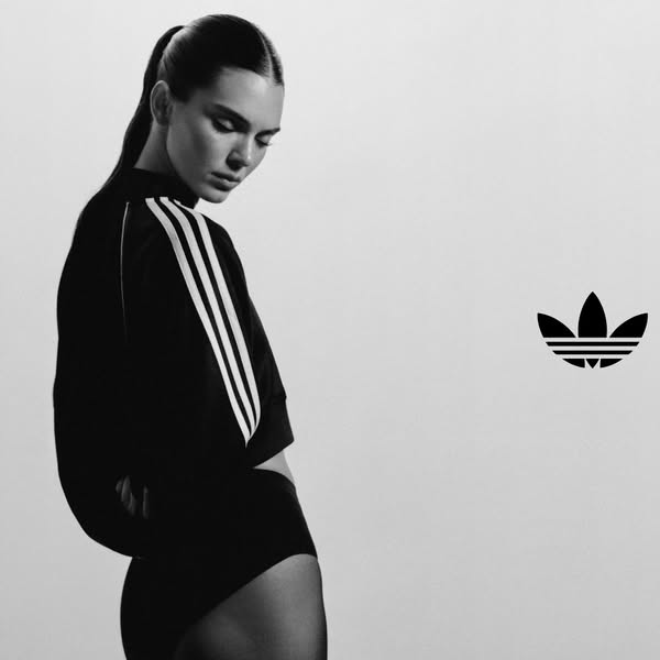 A black and white ad for Adidas. The ad features a model wearing a black long-sleeved cropped shirt with the three white