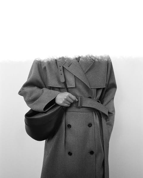 This black and white image ad features a woman wearing a trench coat and carrying a black bag. The ad focuses on the bag