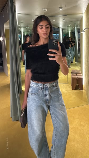 A woman models a fuzzy black top and loose fitting jeans while taking a selfie in a dressing room with mirrors. The ad h