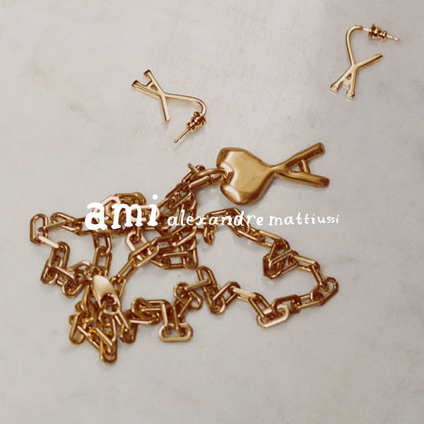 This ad showcases Ami Alexandre Mattiussi jewelry, including earrings and a necklace, arranged on a textured surface.