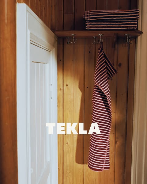 This image ad showcases the TEKLA brand and one of its striped bath towels hung on a hook in a bathroom. 