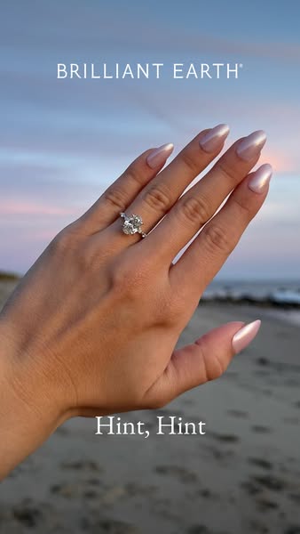 This ad features a close-up of a hand wearing a Brilliant Earth engagement ring on a beach at sunset. The text "Hin