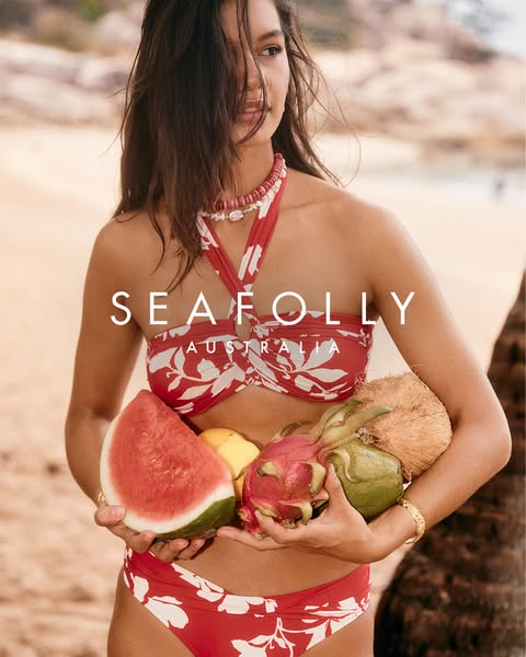 This ad showcases a woman on a beach, wearing a red and white floral print bikini from Seafolly Australia. She is holdin