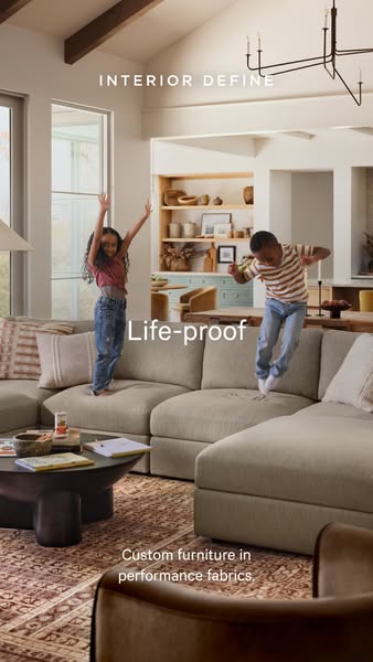 This image ad for Interior Define features two children jumping on a large sectional sofa in a well-lit living room. The