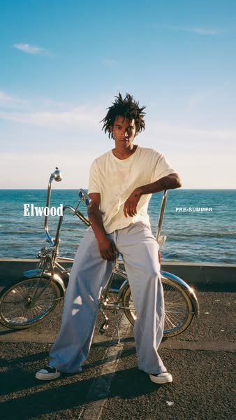 This image ad features a model sitting on a bicycle in front of a beach scene, promoting Elwood clothing.