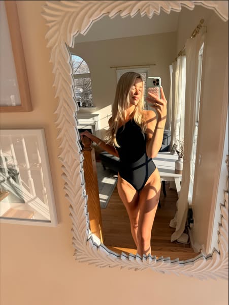 The image is a selfie taken in a mirror with the subject wearing a black one-piece swimsuit. The overall feel is casual 