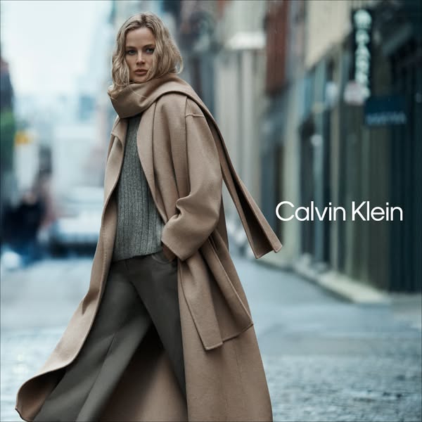 An image ad from Calvin Klein showing a blonde woman wearing a long tan overcoat and walking down a city street.