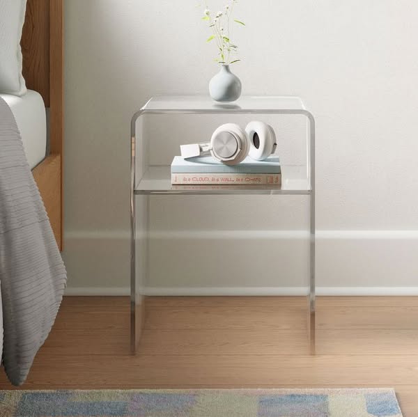 This ad showcases a clear acrylic side table in a staged bedroom setting. The table is styled with headphones and books,