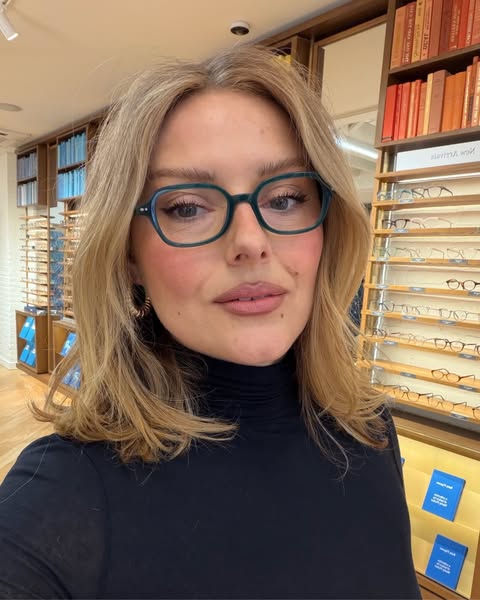 A woman is wearing a pair of glasses while she is in an eyewear store. Shelves filled with books and eyewear can be seen