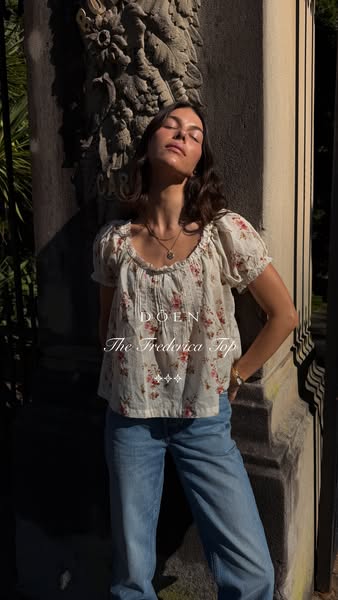 A woman models a floral print top from the brand DOEN. She is standing in front of a stone wall, leaning against a carve