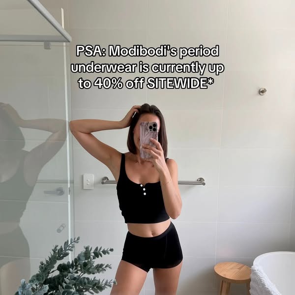 This image ad features a woman in a black Modibodi top and underwear set taking a mirror selfie in a bathroom. The ad pr