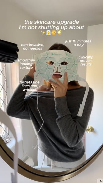 A woman is standing in front of a mirror holding a light therapy mask up to her face. On screen text and arrows call out