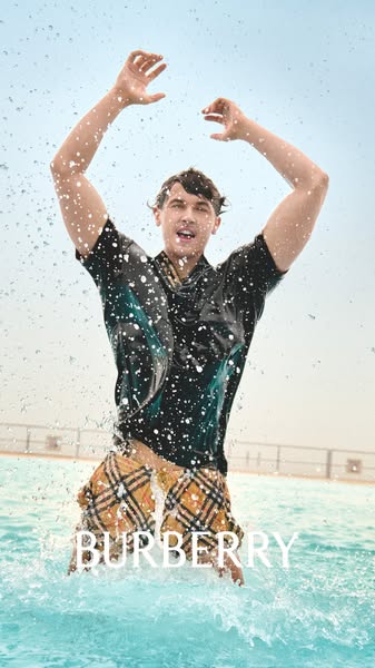 This image is a Burberry ad featuring a man standing in water, wearing Burberry swim trunks and a top. The image showcas