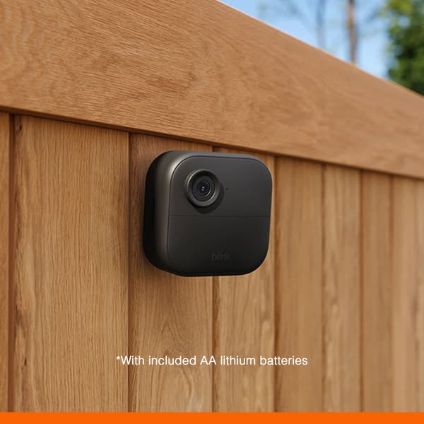 A close up image of a Blink security camera mounted on a wooden wall. The image shows the sleek, unobtrusive design of t