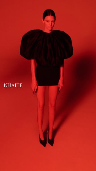 This ad for Khaite features a model in a black dress in a starkly lit red room. The dress has a large, exaggerated top, 