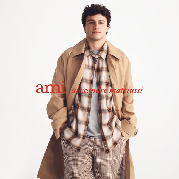 This image features a man modeling Ami clothing. He wears a tan trench coat, a plaid button-down shirt, a gray t-shirt,