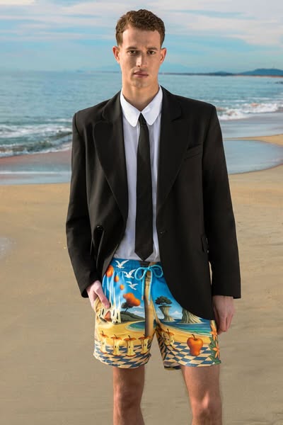 A man in a business suit top with swim trunks on a beach.