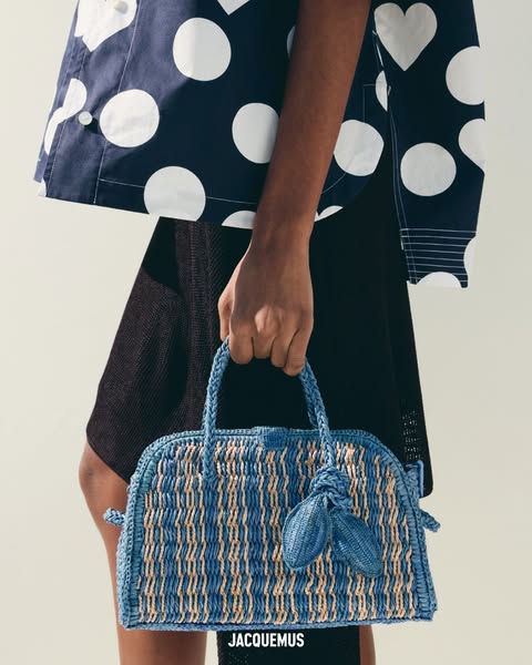 A product shot showcases a blue straw bag from the brand Jacquemus. The bag is held by a person who is wearing a navy an