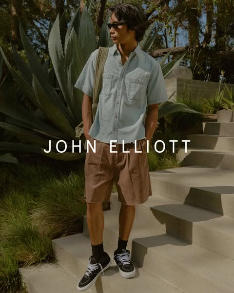This ad showcases a person wearing John Elliott brand shorts and a short-sleeve button-up shirt in an outdoor setting.