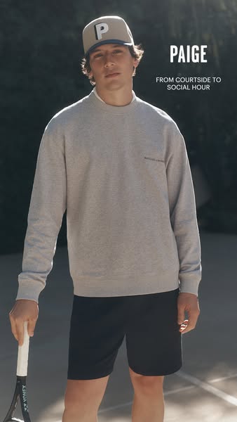 A male model stands on a tennis court holding a tennis racket wearing apparel by Paige.