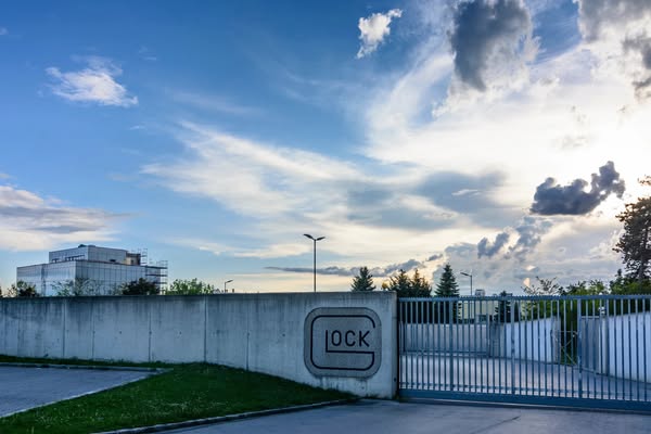 This image ad showcases the exterior of a Glock factory, set against a backdrop of a cloudy sky and a modern office buil