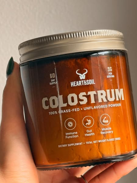 An image ad for Heart & Soil colostrum. The image shows a hand holding a clear jar of colostrum.