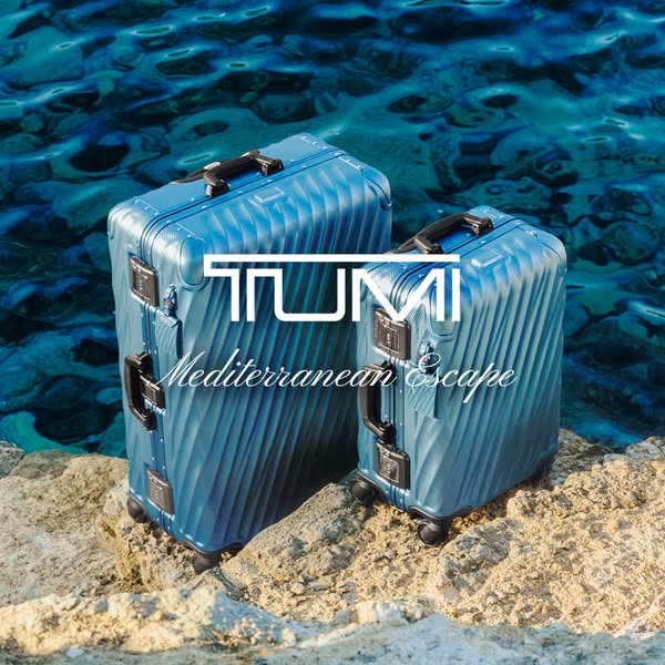 This ad for Tumi luggage features two blue metallic suitcases on a rocky surface, set against a backdrop of blue ocean w
