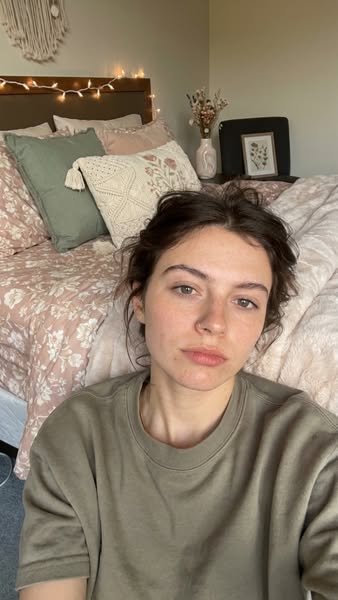 A woman lies on a bed and looks directly at the camera in selfie style. There are pillows and a comforter behind her and