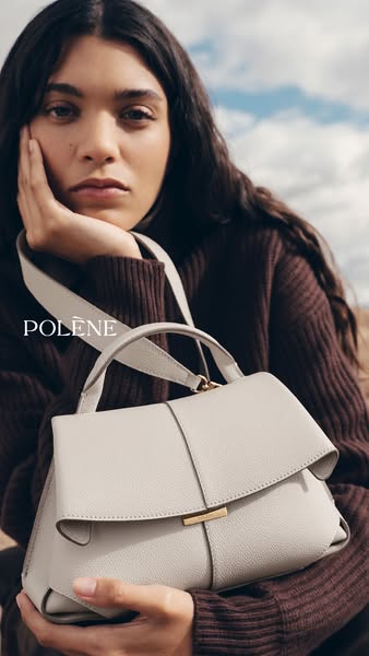 This image ad showcases a Polène bag. A person is holding the cream-colored bag while posing outside with a sky and fiel