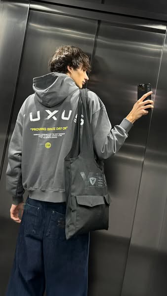 A person stands in an elevator and is holding a phone and taking a mirror selfie. They are wearing clothing and a bag fr