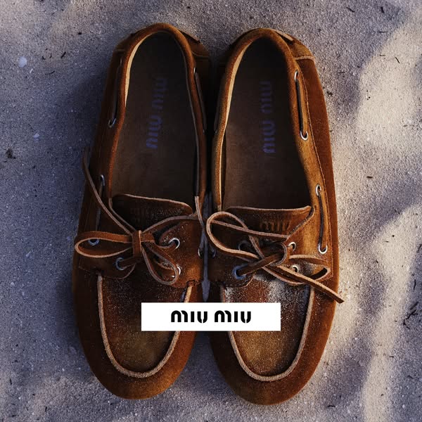 This image ad showcases brown Miu Miu boat shoes on a sandy surface, emphasizing the brand's logo.