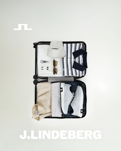 An overhead shot of a suitcase packed with J.Lindeberg branded clothing and accessories, all in neutral tones. The suitc