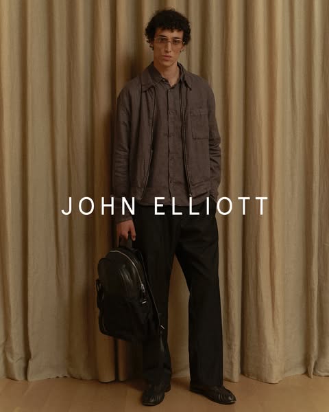 An image ad for John Elliott featuring a person standing with a bag and wearing a jacket and pants from John Elliott'