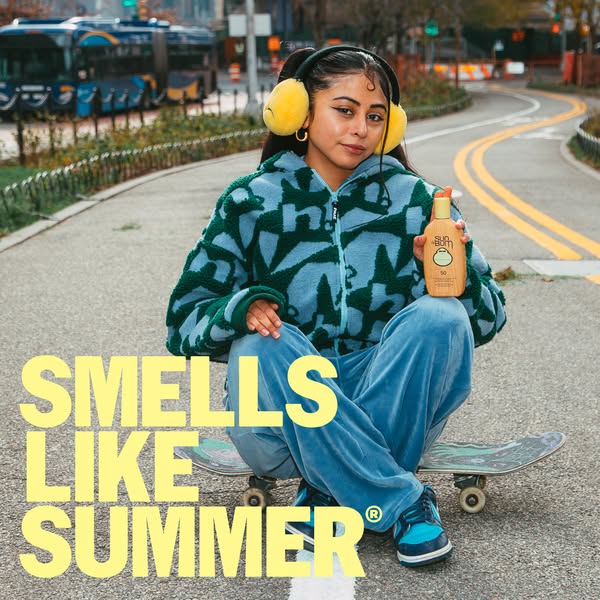 An image ad featuring a person sitting on a skateboard in a park, holding a bottle of Sun Bum sunscreen. The text "