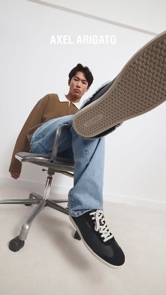 A person sits in a chair with their leg up, showing the bottom of their shoe to the camera. The brand is displayed at th