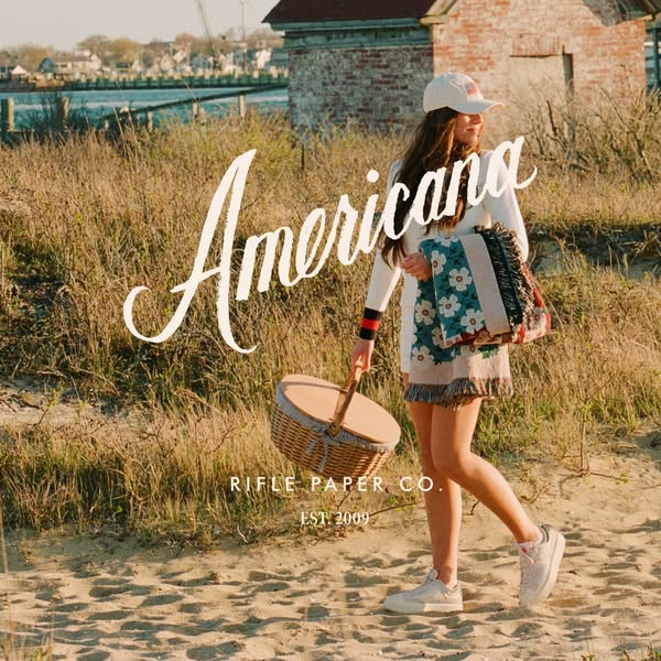 This ad for Rifle Paper Co. features a young woman walking on a beach with a picnic basket and a tote bag. The image has