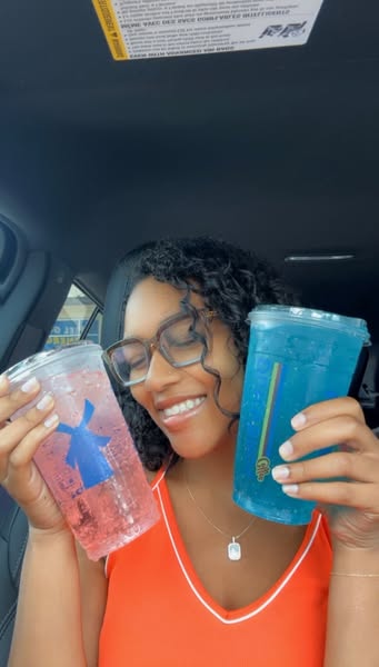 A woman is smiling while holding two large plastic cups filled with colorful beverages from a convenience store. She is 