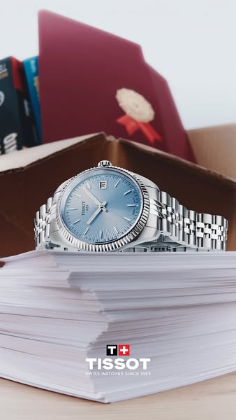 This ad showcases a silver and light blue Tissot watch, lying on top of a stack of papers in front of an open cardboard 