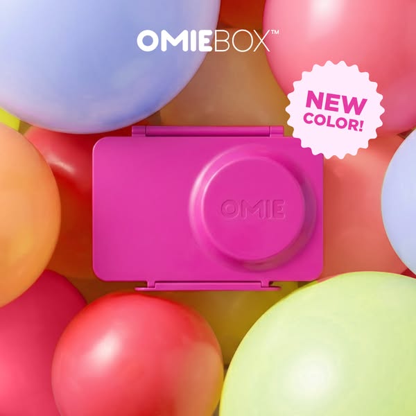 This image ad shows a pink OmieBox lunchbox against a background of colorful balloons. The ad announces the new color fo
