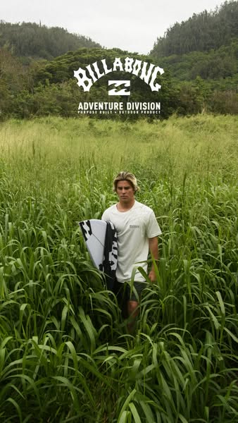 A man is standing in a field of tall grass carrying a surfboard. The image promotes Billabong Adventure Division apparel
