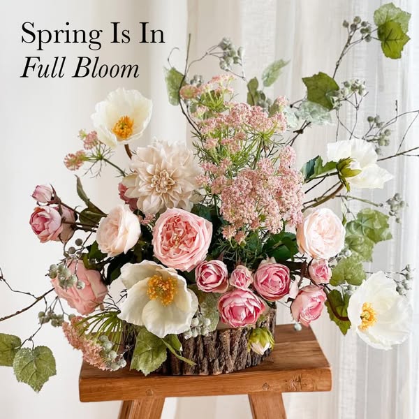 This image ad features a spring floral arrangement in a wood-look vase set on a small wooden table in front of white cur