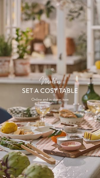 An image ad showcasing a curated tablescape with various serving dishes, a candle, drinks, and fresh produce, emphasizin