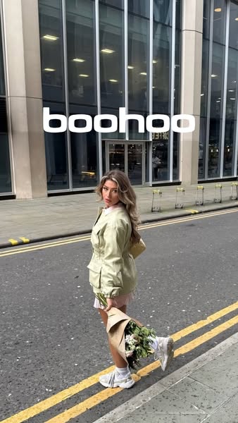 A woman stands on a sidewalk in front of a Boohoo store. She is holding flowers.