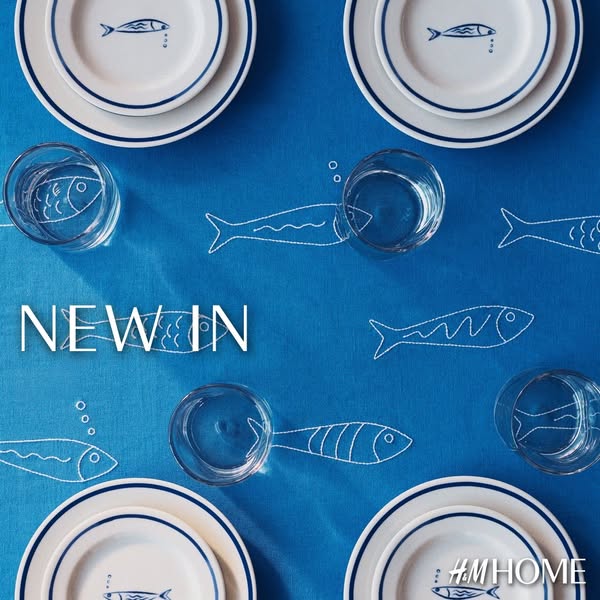 The ad features a flat lay of H&M Home products, including plates and glasses, on a blue tablecloth with white fish 