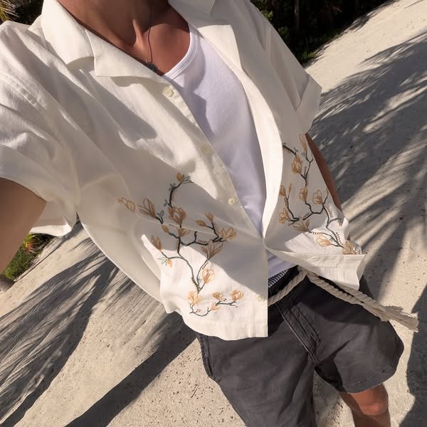 An image of a person wearing a white button-down shirt with floral embroidery paired with dark shorts and a rope belt.