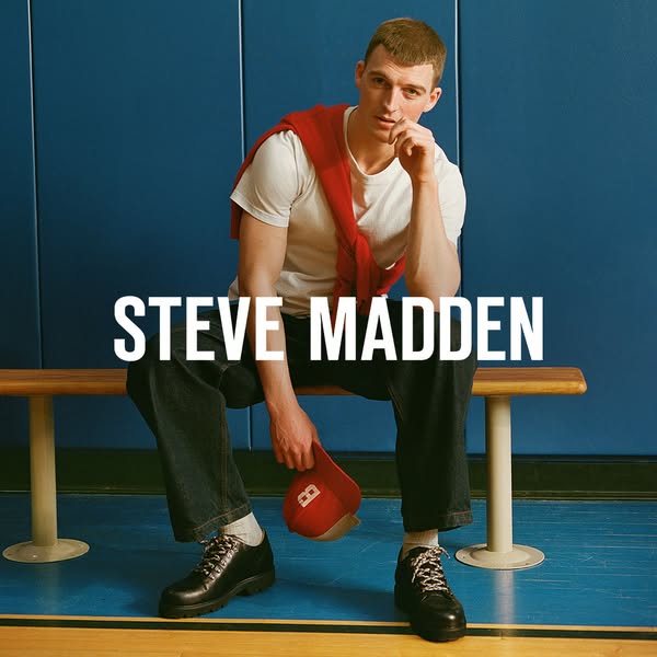 This Steve Madden ad features a man sitting on a bench in front of a blue wall wearing a white t-shirt, jeans and black 