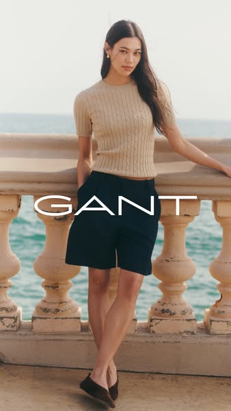 This ad features a woman in a sand-colored cable knit sweater, blue shorts, and brown shoes, leaning against a cream-col
